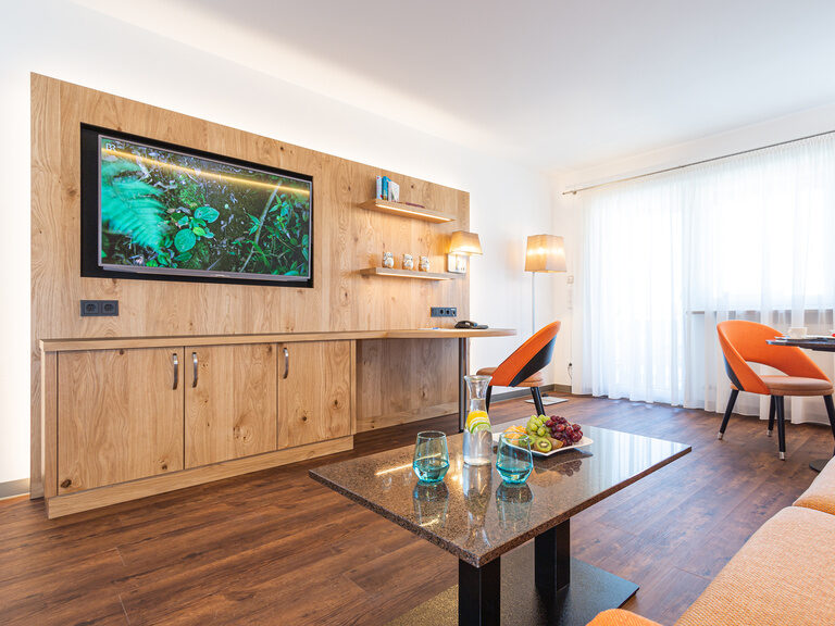 Aufnahme-stillvoller-wohnbereich-2-raum-suiten stillvoller-wohnbereich-mit-vinylboden-in-neuer-2-raum-suite-mit-heller-holz-tvwand-orangen-sesseln-und-schlafcouch-gedeckter-couchtisch-sowie-esstisch